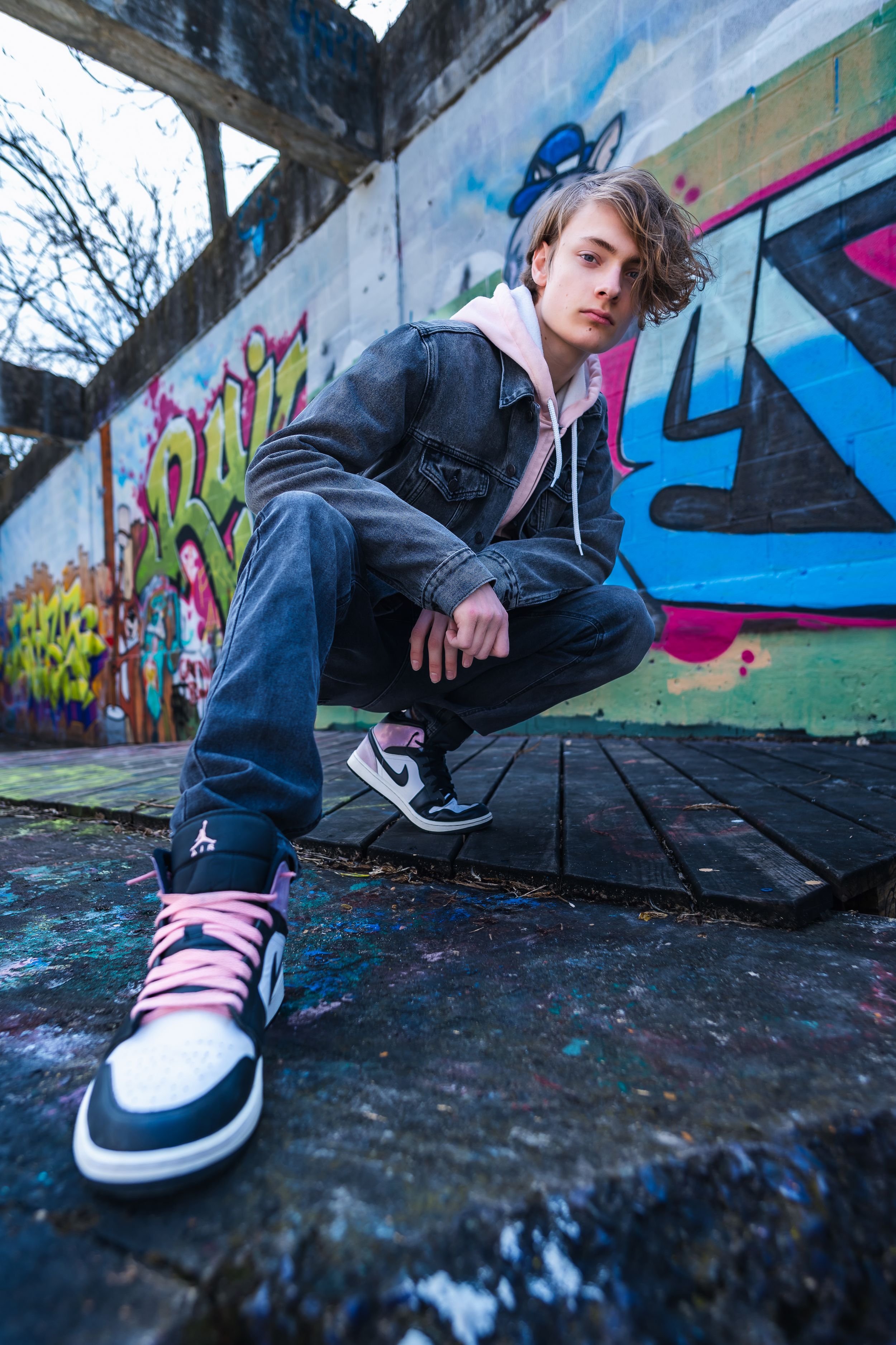 Teen boy in denim jacket and pink Jordan sneakers crouching by graffiti wall.