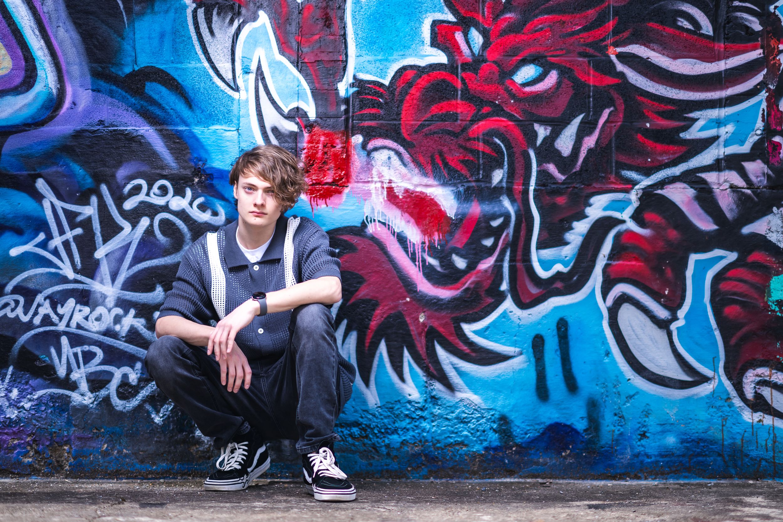 Young man sitting against a vibrant blue wall with red dragon graffiti street art in urban setting.