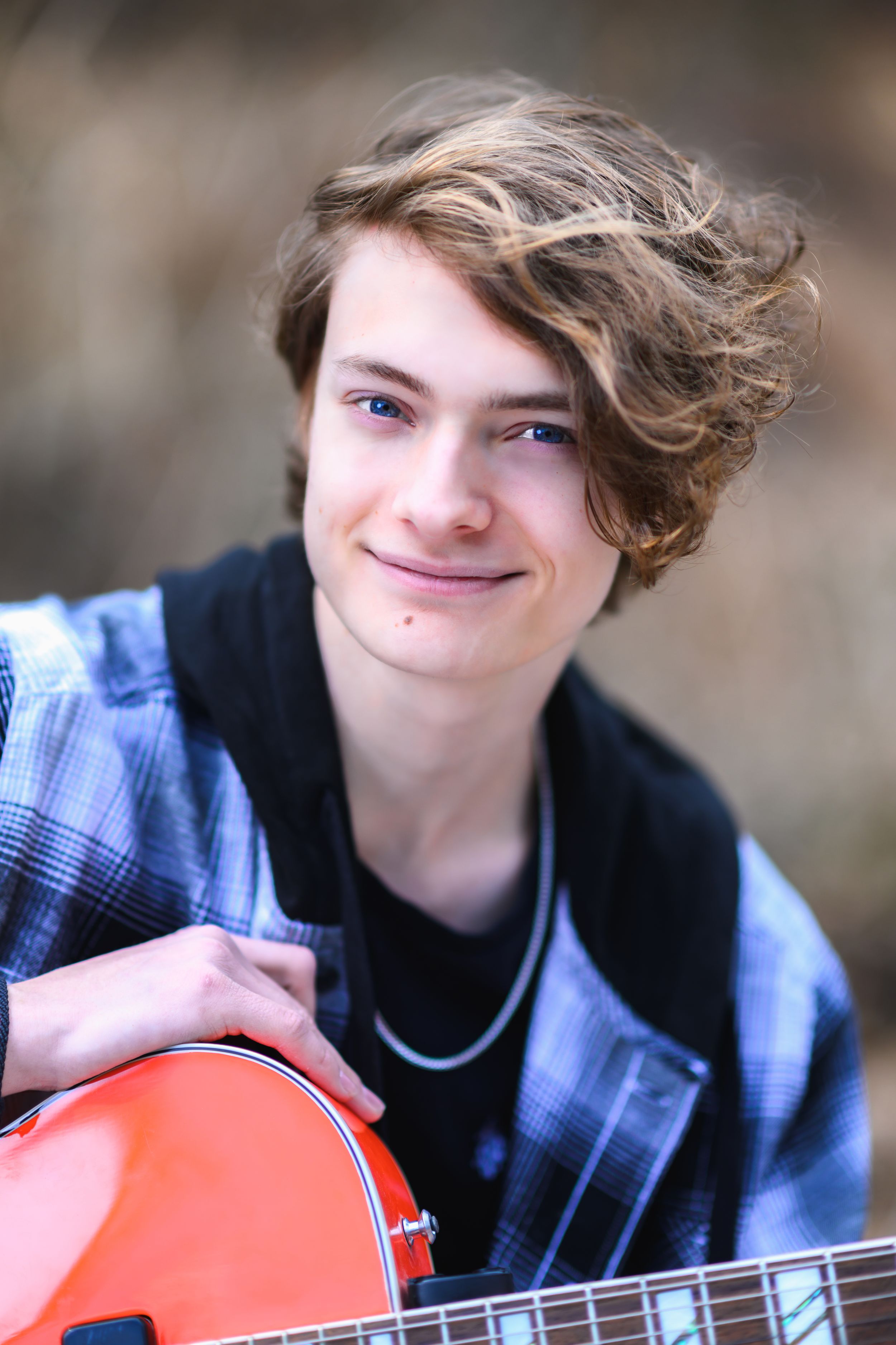 Smiling teen boy with wavy hair holding a red electric guitar outdoors.