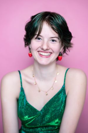 A person in a green sparkly dress poses against a pink background wearing red earrings.