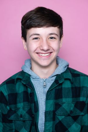 A person in a plaid flannel and grey hoodie smiles against a pink background.
