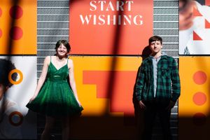A couple poses against a vibrant orange wall with a 'START WISHING' sign in an artistic photoshoot.