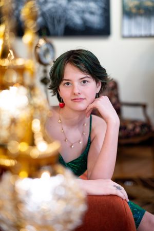 Portrait in emerald green dress with red earrings beside golden chandelier lighting.