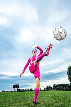 A female soccer goalkeeper in pink uniform skillfully juggles a soccer ball on a green field under a cloudy sky.