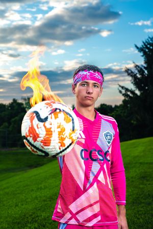 Teen soccer player in pink jersey holding a flaming ball on a field.