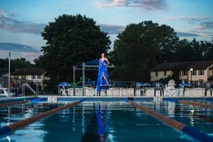 A woman in an elegant blue gown stands gracefully on a diving board above a reflective outdoor pool at dusk.