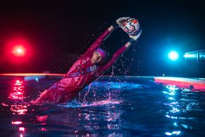Athlete in red suit catches ball in illuminated pool with dramatic blue and red lighting at night.