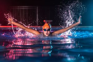 A competitive swimmer performs the butterfly stroke in a pool illuminated by dramatic pink and blue neon lights.