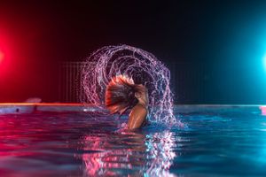 Woman flipping wet hair in a pool, creating a water arc under dramatic red and blue neon lights.
