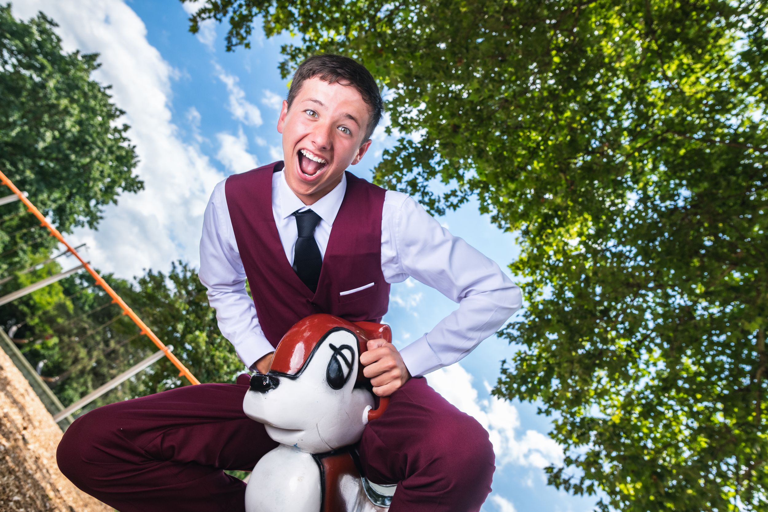 Excited young man in maroon vest poses with cartoon dog statue outdoors under blue sky and green trees.