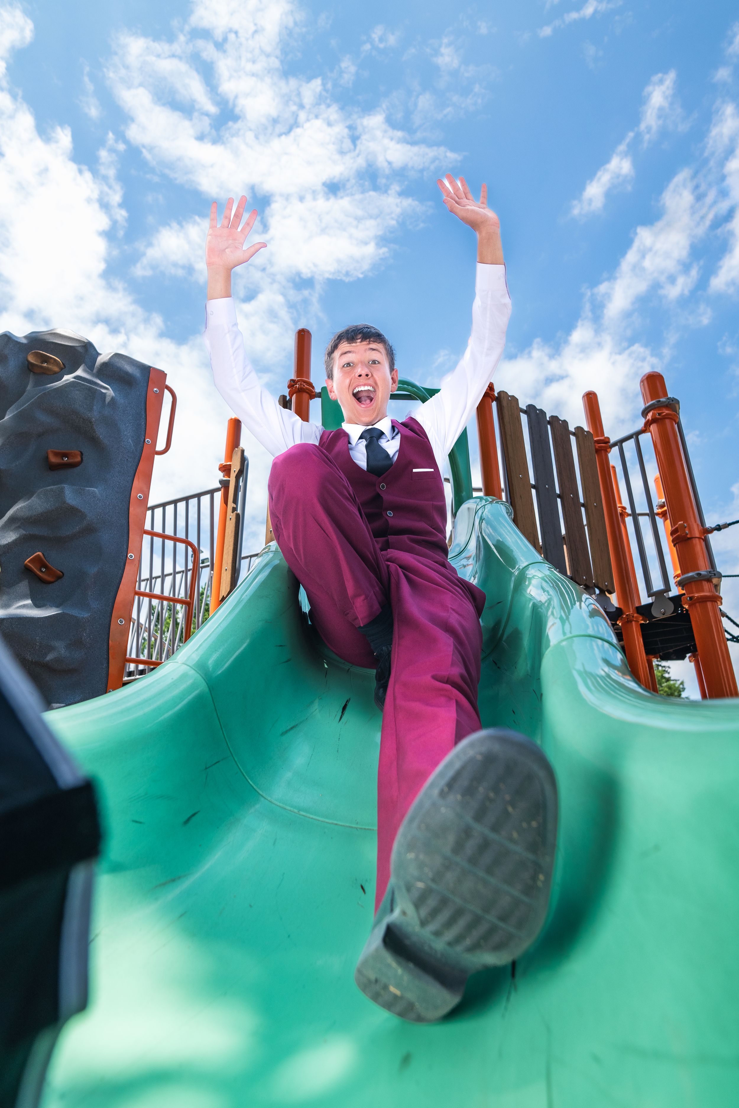 Boy in purple suit sliding down playground slide with arms raised in excitement under blue sky.