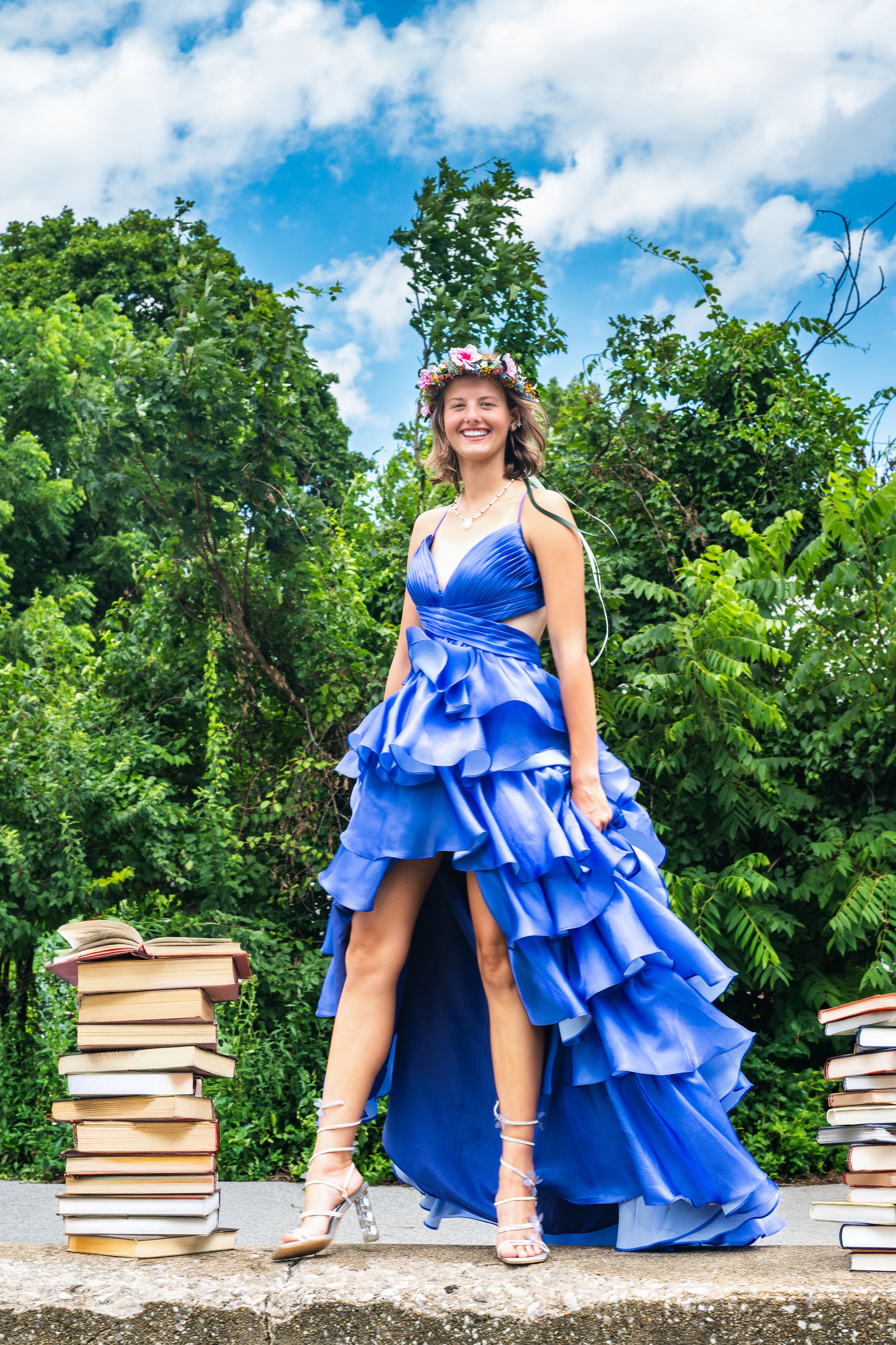 Woman in blue ruffled gown and floral crown stands between stacks of books outdoors.