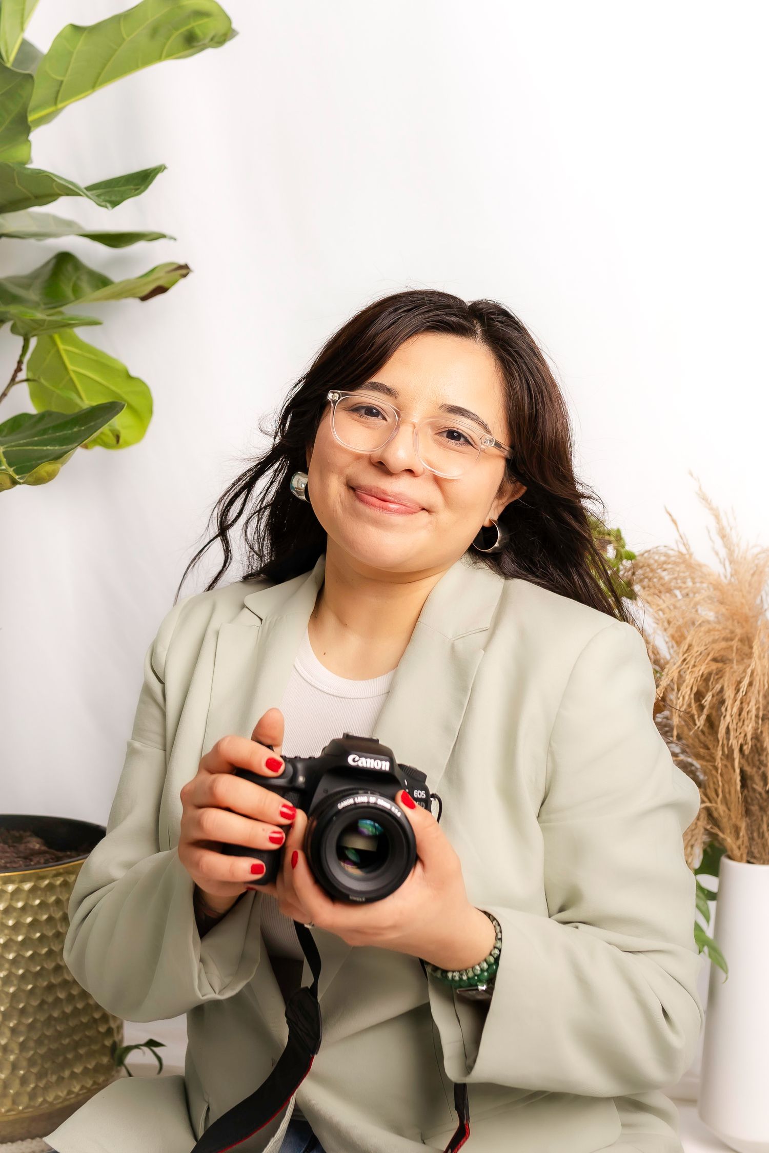 Smiling photographer in sage blazer holding camera against white wall with plants.
