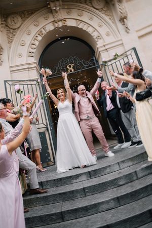 Newlyweds celebrate joyfully on church steps with wedding guests cheering and throwing flower petals.