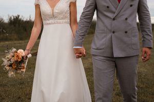 Newlywed couple holding hands outdoors with bride in white dress holding peach bouquet and groom in grey suit.