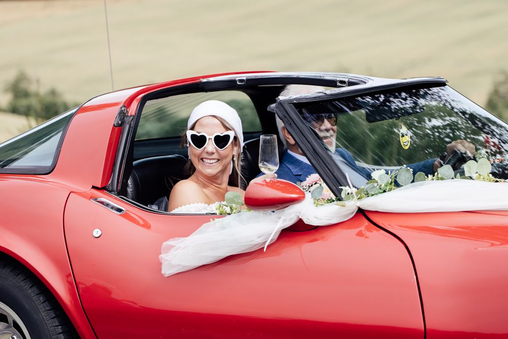 Bride in heart sunglasses smiling in a decorated red convertible sports car with groom on wedding day.