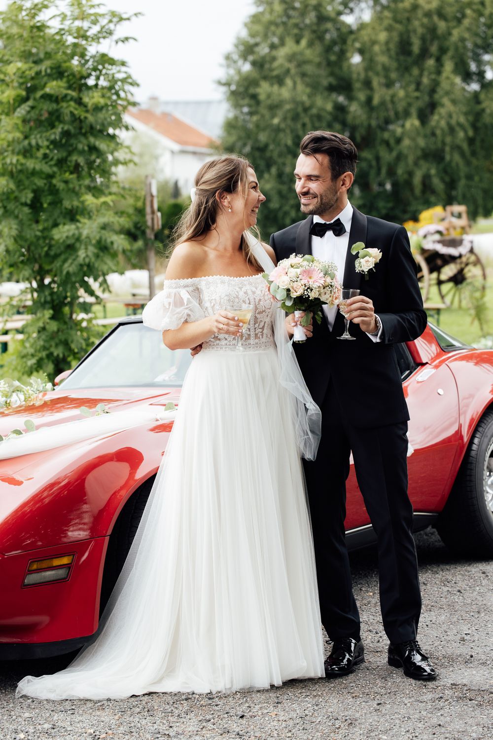 Bride and groom holding champagne glasses stand beside a red convertible sports car on their wedding day.
