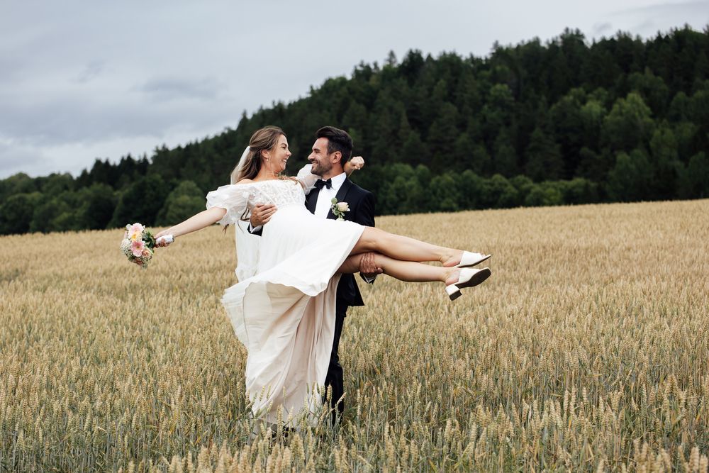 Groom lifting bride in white dress in golden wheat field with forest backdrop on their wedding day.