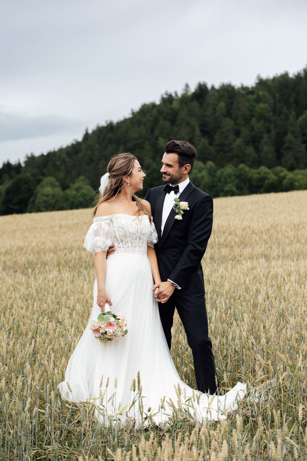 Bride and groom holding hands in a golden wheat field, wearing white gown and black tuxedo, with forest backdrop.