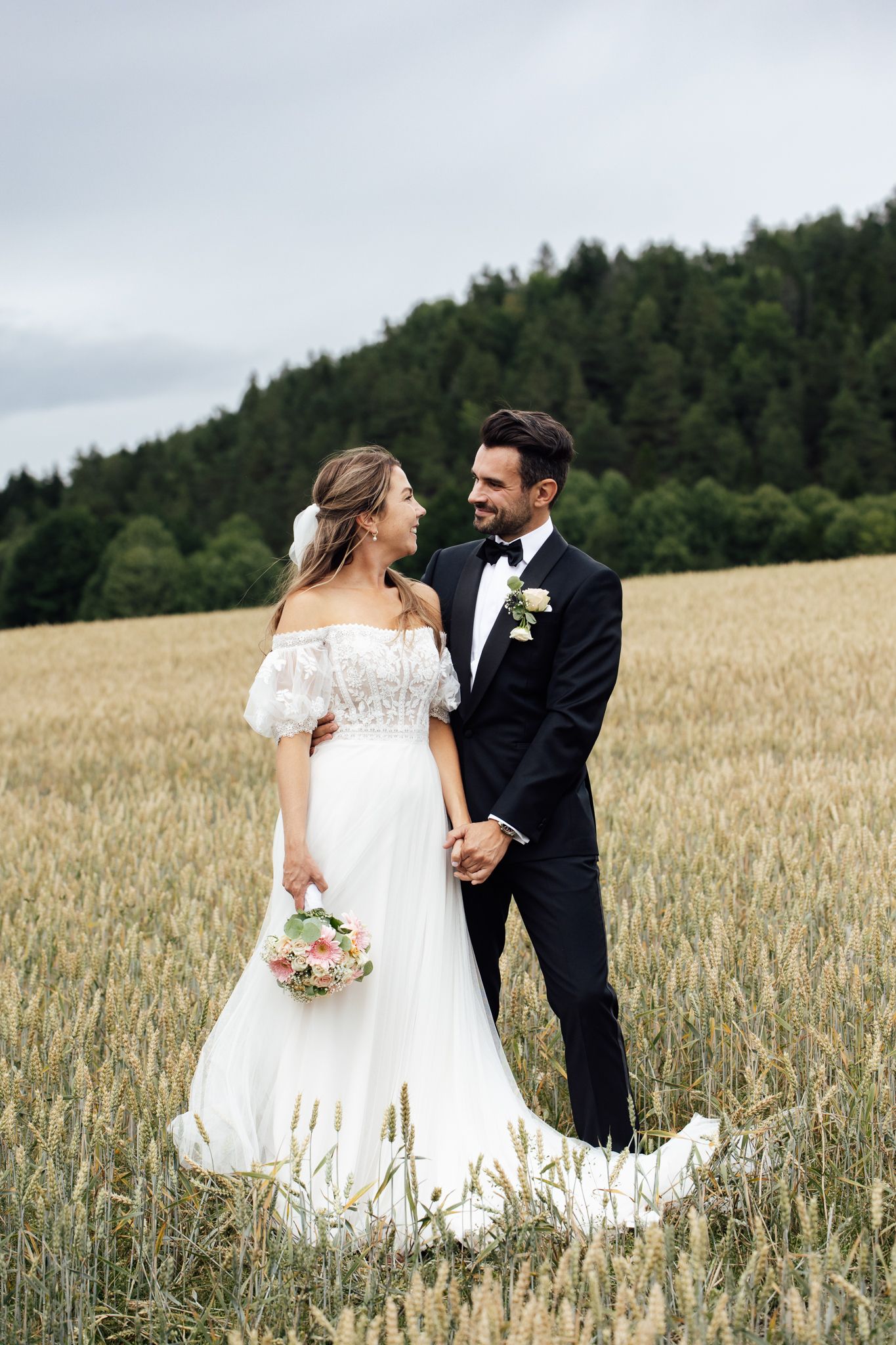 Bride and groom holding hands in a golden wheat field, wearing white gown and black tuxedo, with forest backdrop.
