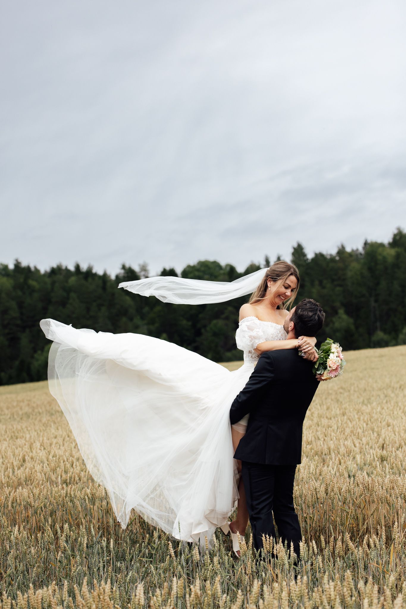 Groom lifting bride in white gown and flowing veil in a golden wheat field on their wedding day.
