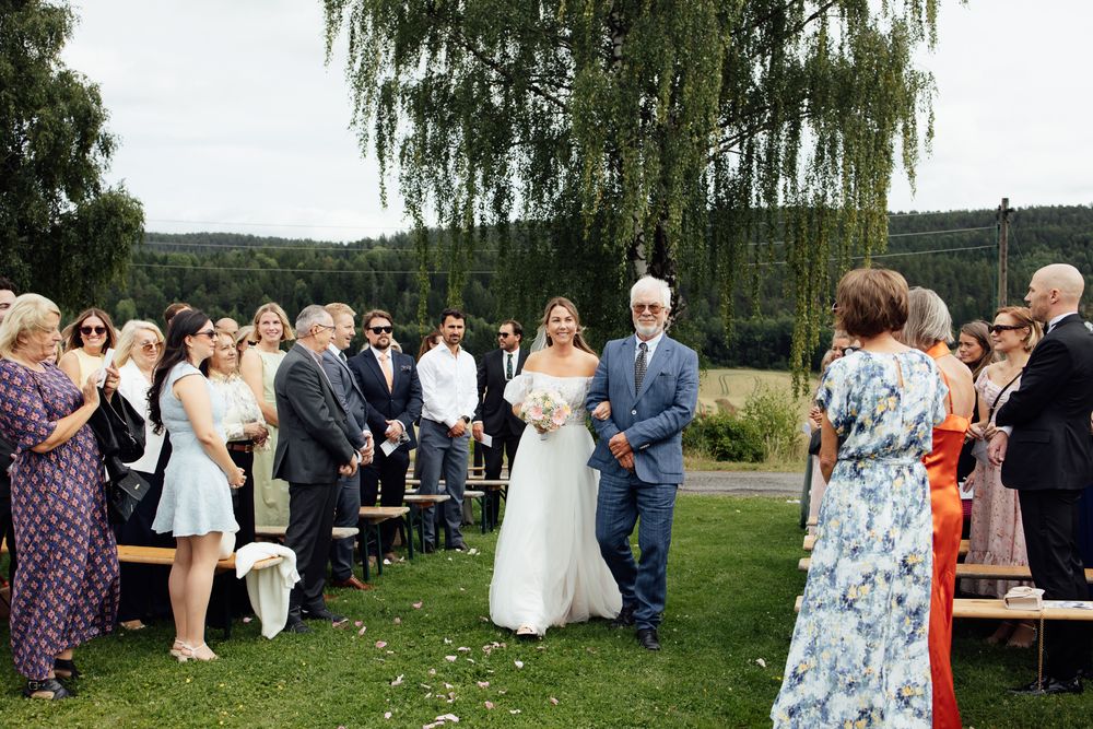 Bride walks down outdoor aisle with escort at garden wedding ceremony, guests seated on benches amid lush green scenery.
