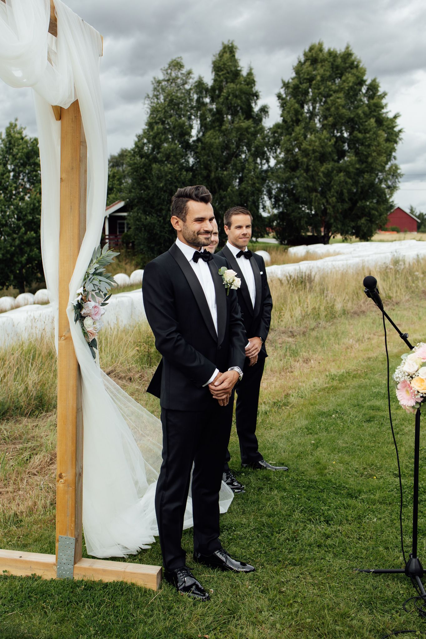 Two grooms in black tuxedos stand at an outdoor wedding altar decorated with white draping and flowers.