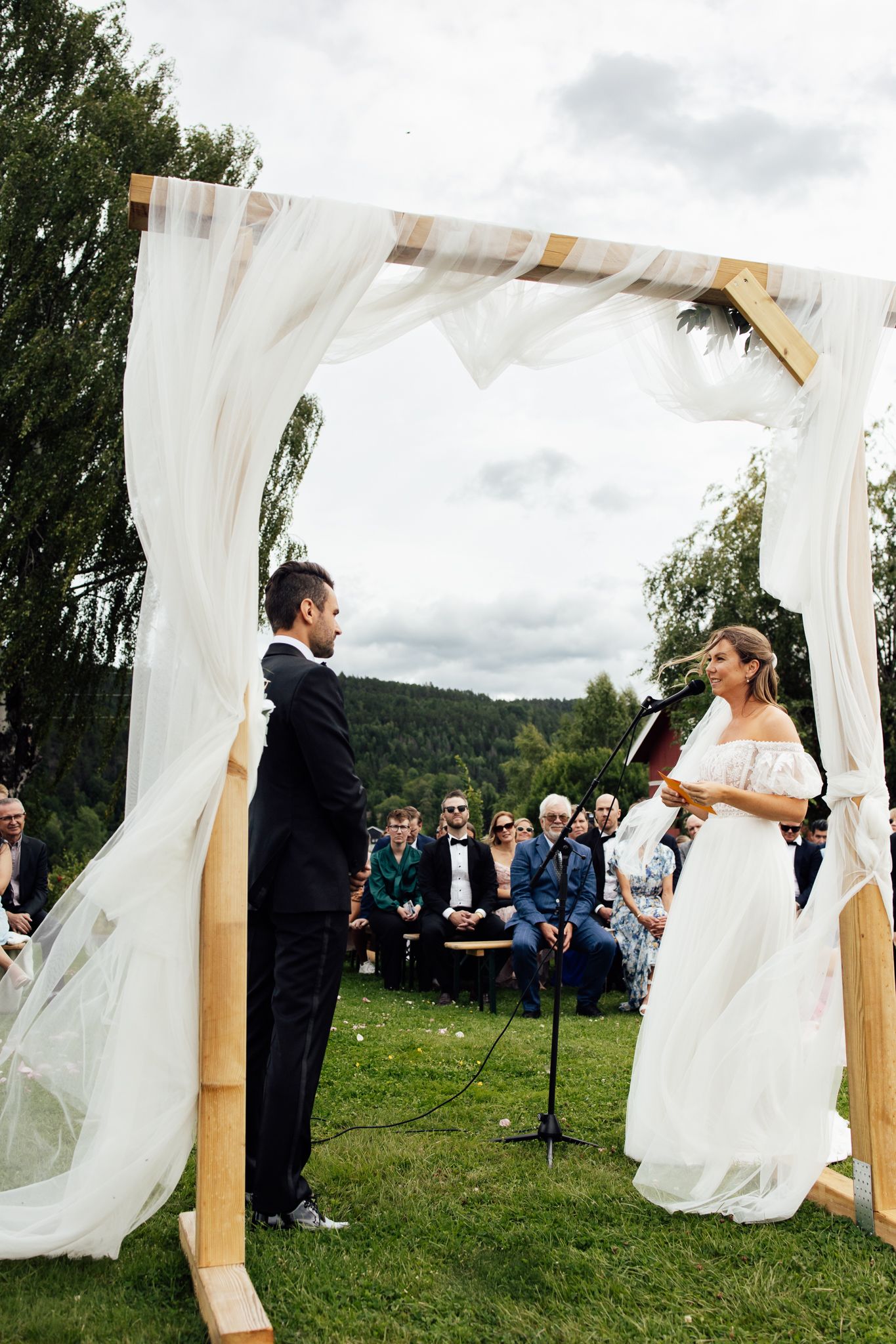 Outdoor wedding ceremony under a wooden arch with white draping, bride in off-shoulder gown facing groom in black suit.