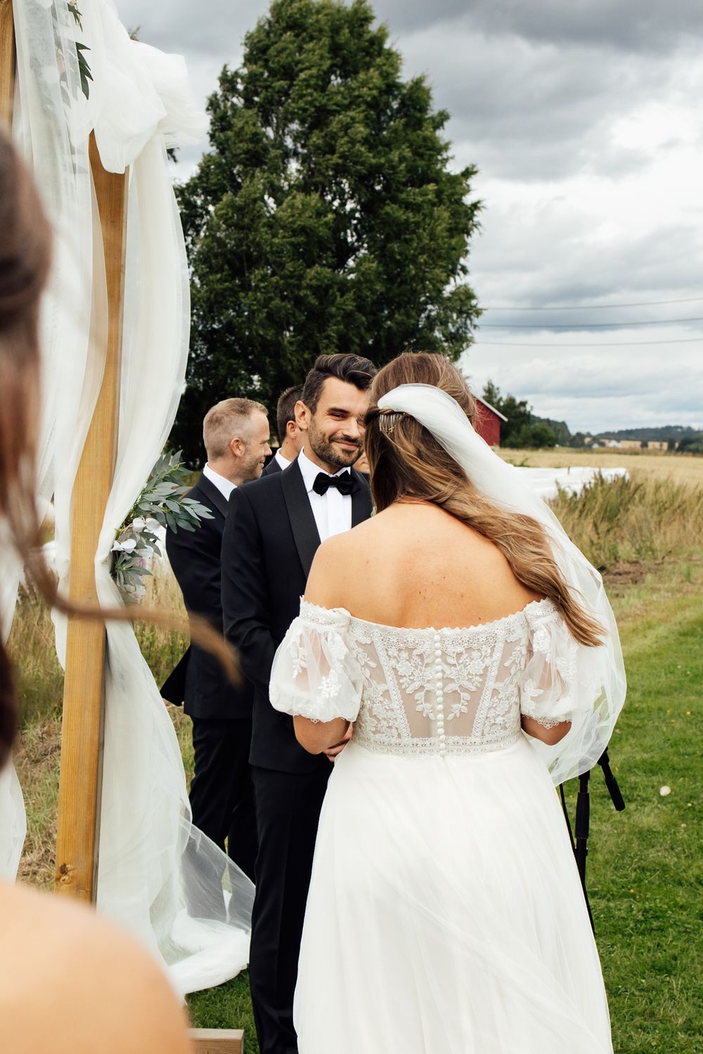 Smiling groom in black tuxedo holds hands with bride in lace off-shoulder wedding dress during outdoor ceremony.