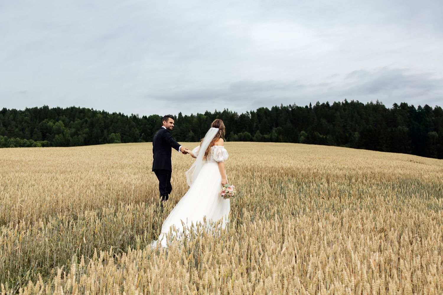 Bride and groom walking hand in hand through a golden wheat field with a forest backdrop on their wedding day.