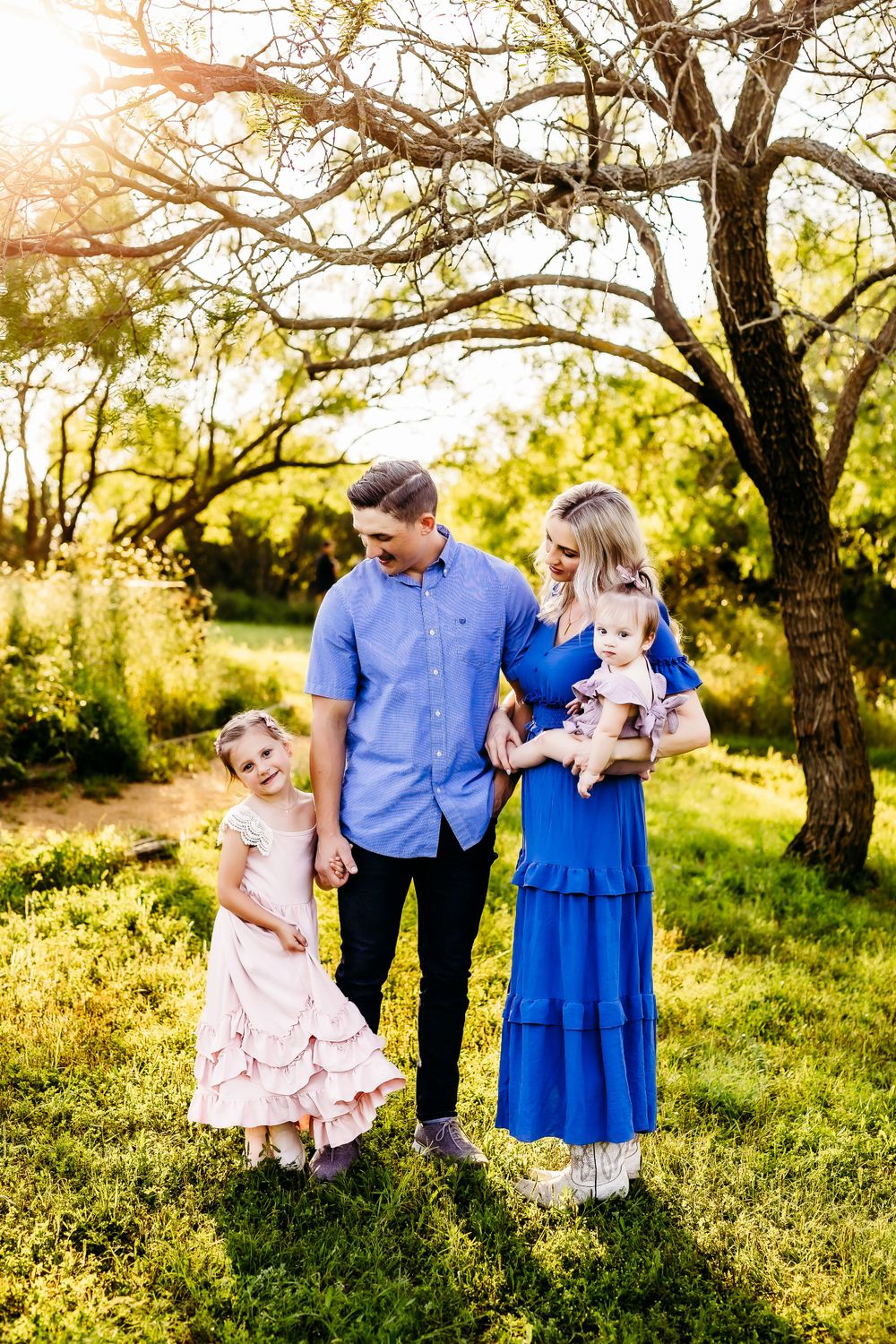 Family portrait in a sun-dappled meadow with blooming trees in the background.
