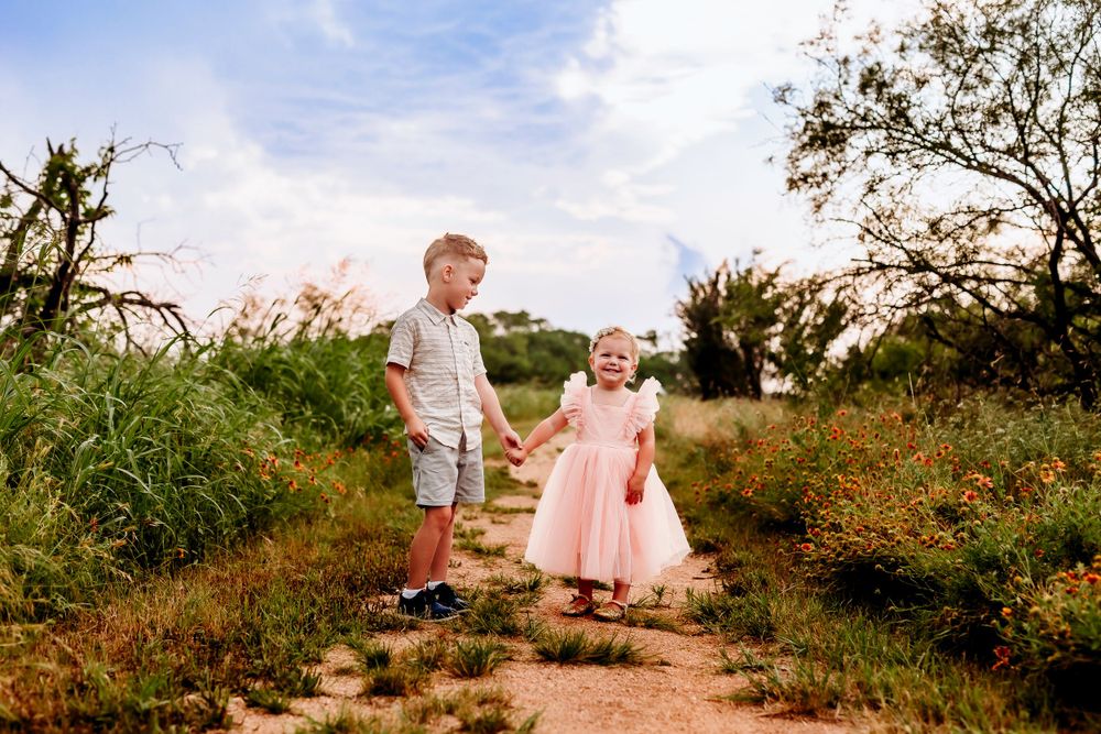 Two children walking together hand-in-hand on a dirt path at sunset surrounded by wildflowers and natural vegetation.