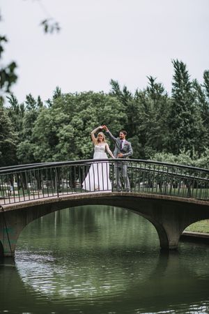 Bride and groom dancing on an arched stone bridge over a calm green pond in a lush park setting.