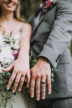Bride and groom showing off wedding rings, holding a white and purple floral bouquet, wearing a gray suit and white dress.