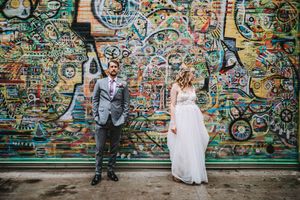 Bride and groom pose holding hands in front of a vibrant colorful graffiti mural wall on their wedding day.