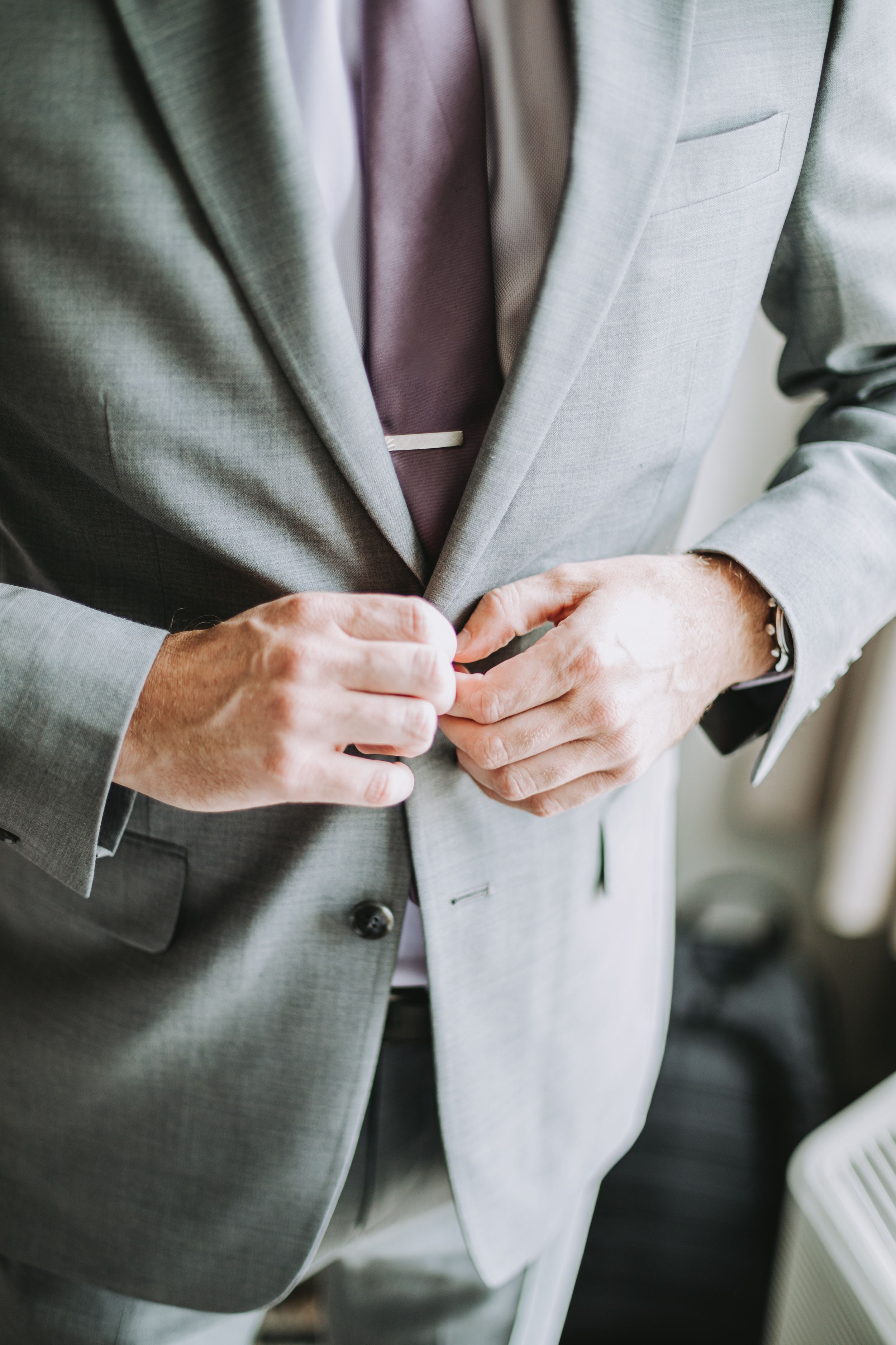 Man in light gray suit buttoning jacket with pink tie and silver watch, formal wedding attire close-up.