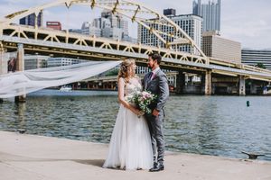 Bride and groom share a romantic moment by Pittsburgh riverfront with iconic yellow bridge and city skyline backdrop.