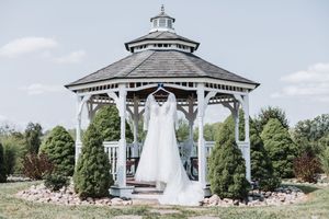 A elegant white wedding dress hanging in a classic white gazebo surrounded by evergreen trees on a sunny day.