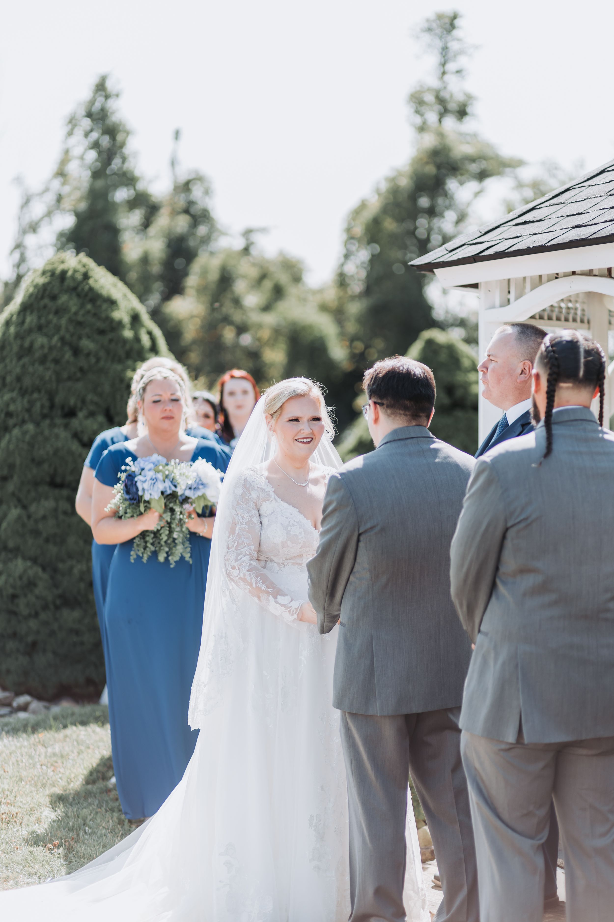 Bride in white lace gown smiles during outdoor wedding ceremony with bridesmaids in blue dresses and groomsmen in gray suits.