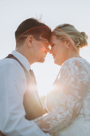 Bride and groom touching foreheads in golden sunset light, sharing a romantic moment in wedding attire.