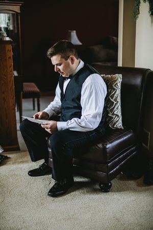 Groom in vest and bow tie reads a letter while seated in a leather chair during wedding day preparations.