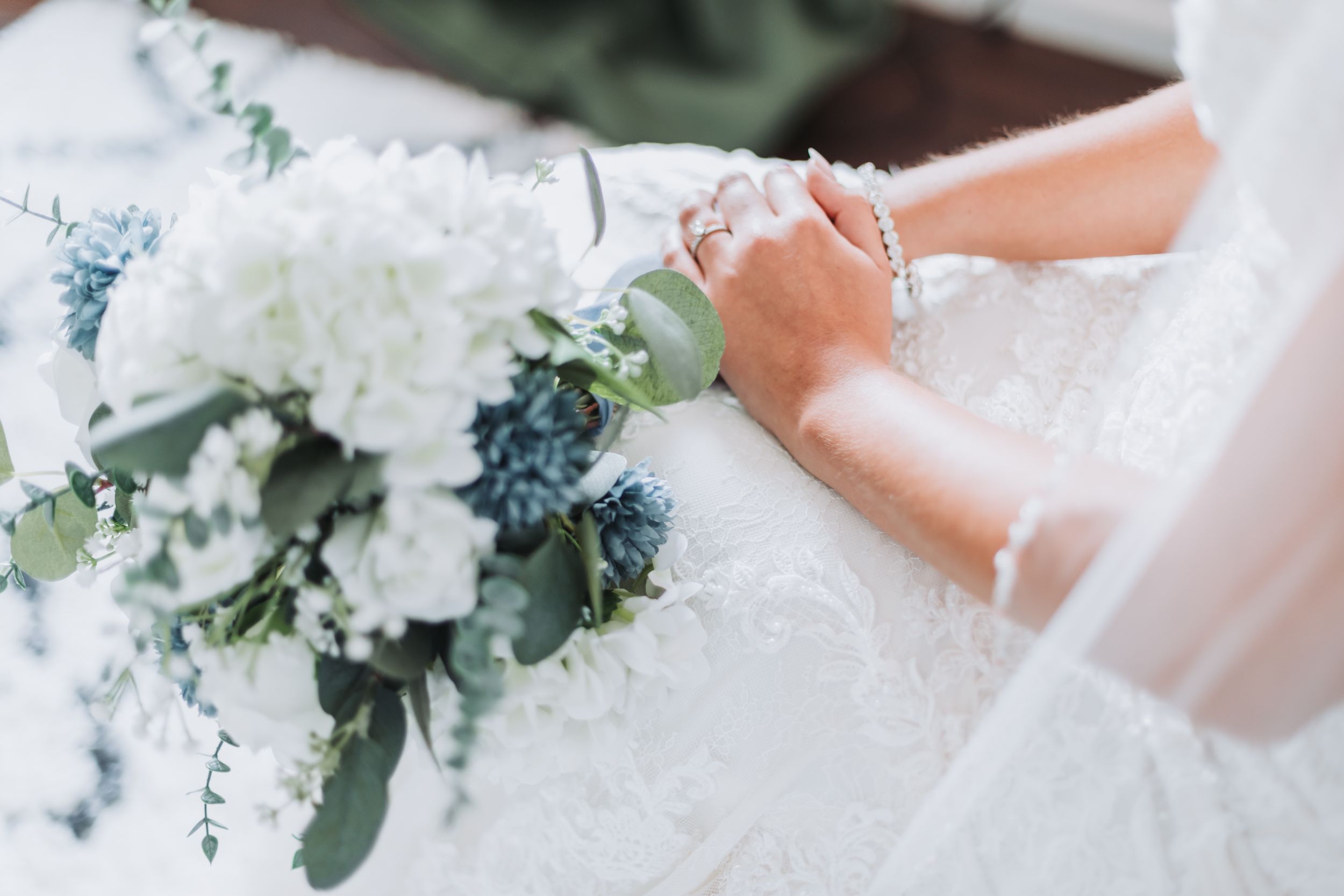 Bride in lace wedding dress holding white hydrangea and blue thistle bouquet with greenery, wearing bracelet and ring.
