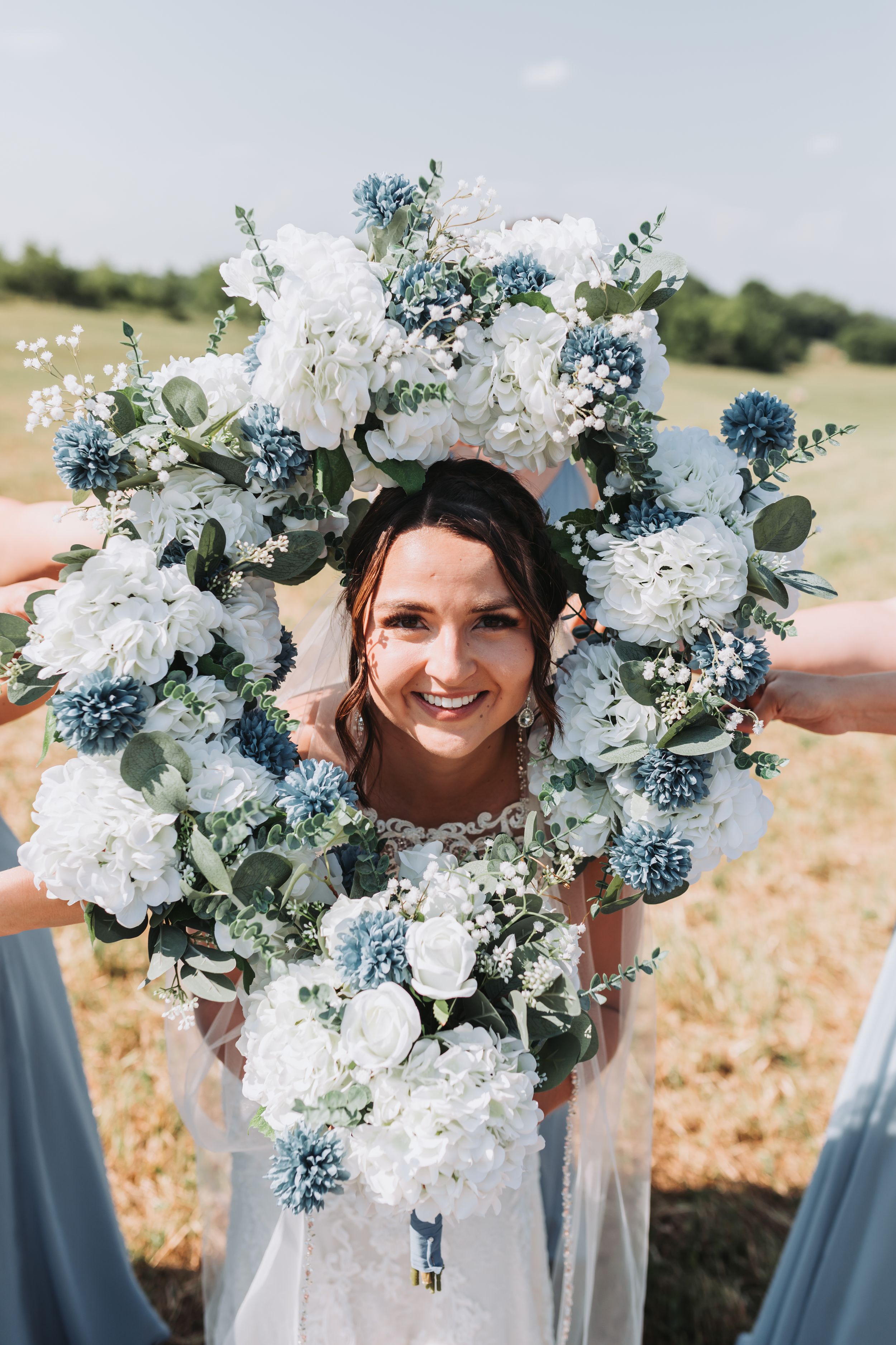 Smiling bride framed by blue and white hydrangea bouquets held by bridesmaids in light blue dresses outdoors.