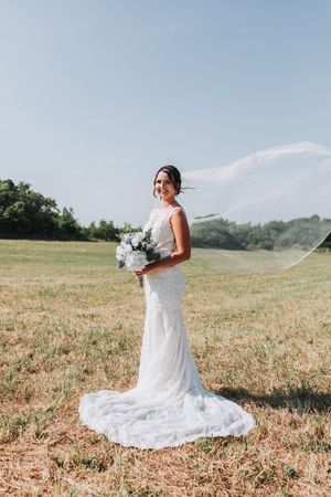 Bride in white lace gown with flowing veil and blue bouquet standing in sunny field on wedding day.