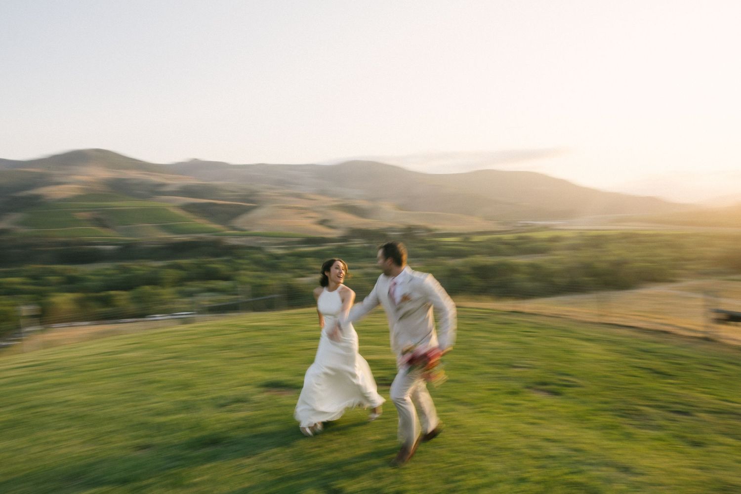 People in white clothing running and dancing across rolling green hills at golden sunset hour in Northern California.