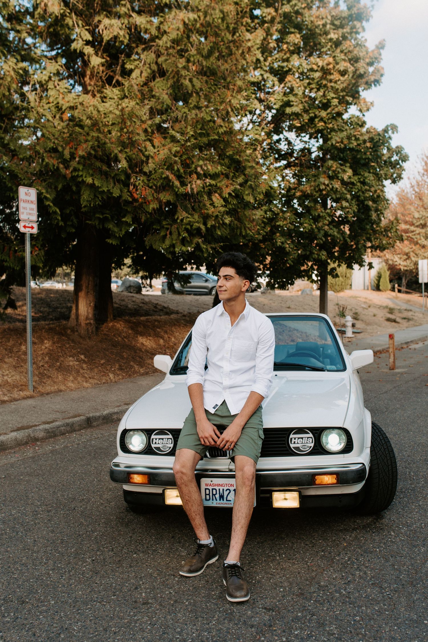 Person sitting on hood of vintage BMW car under fall trees on residential street.