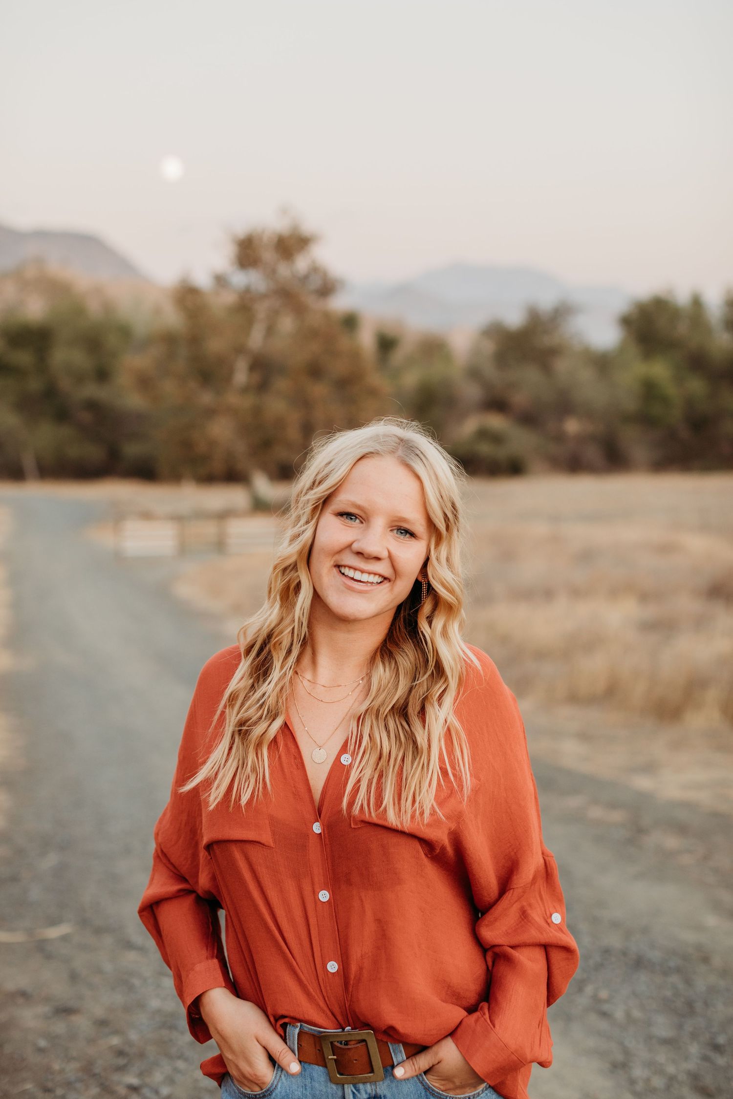 Person in burnt orange button-down shirt poses outdoors against mountain backdrop at sunset.