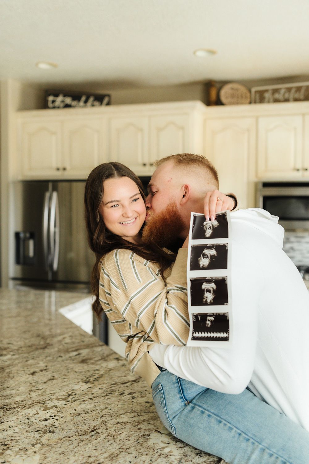 Couple sharing emotional moment with ultrasound photos on marble kitchen counter.
