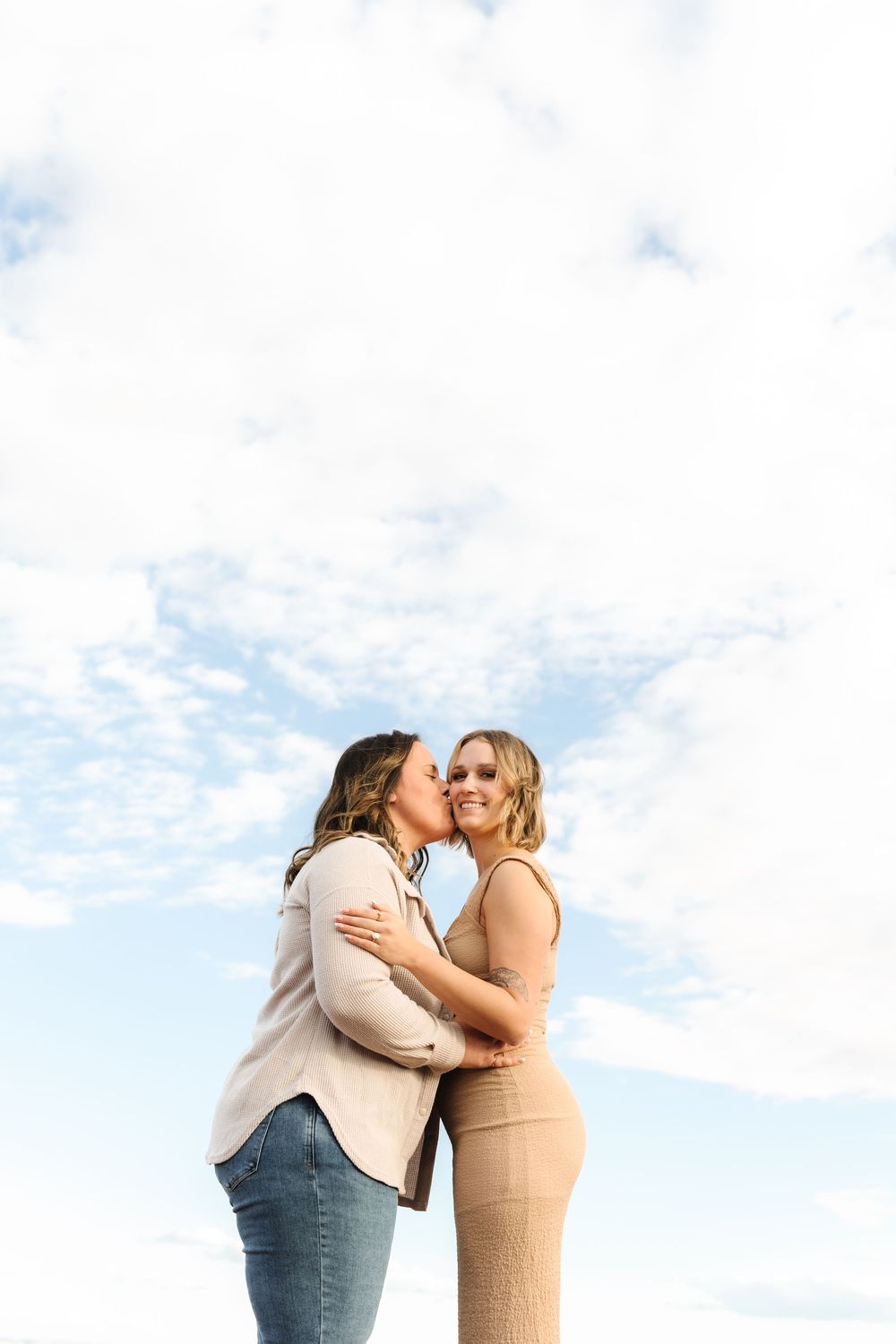 Intimate couple embracing against blue sky with wispy clouds.