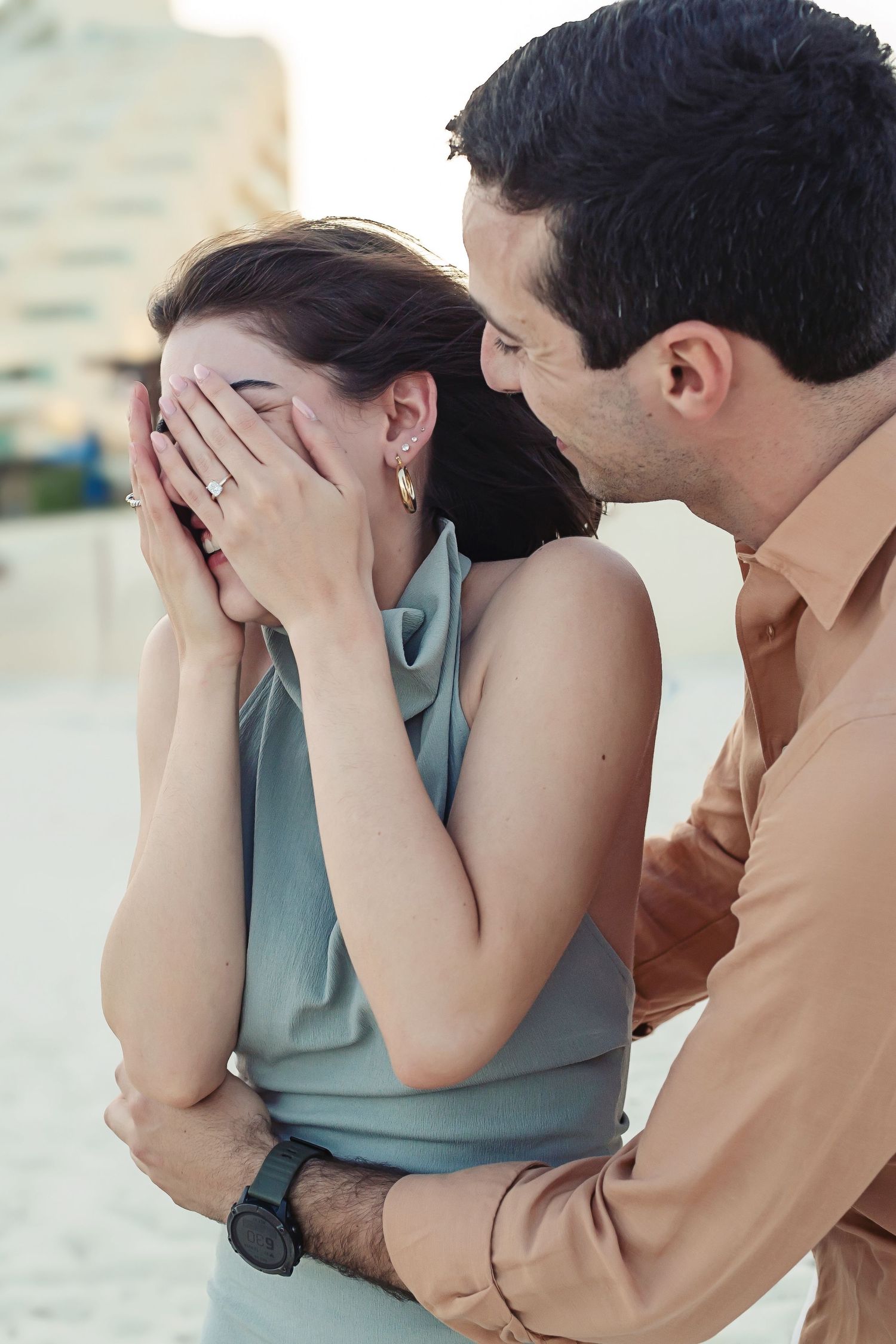 A romantic moment at sunset as a couple embrace, with one person covering their face while the other leans in close.